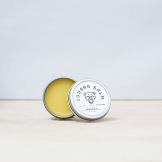 Cougar Balm, a soft pale yellow salve, is featured cap off with the lid leaning against it. 