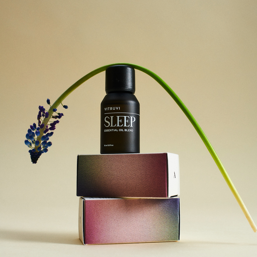 The 'Sleep' essential oil blend sits on top of two boxes. A long stemmed flower is wrapped over the top of the bottle. 